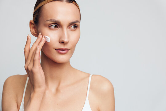 Young woman applying face cream on clean skin, skincare routine, natural beauty, soft lighting, white background, focused expression, casual wear, healthy lifestyle concept