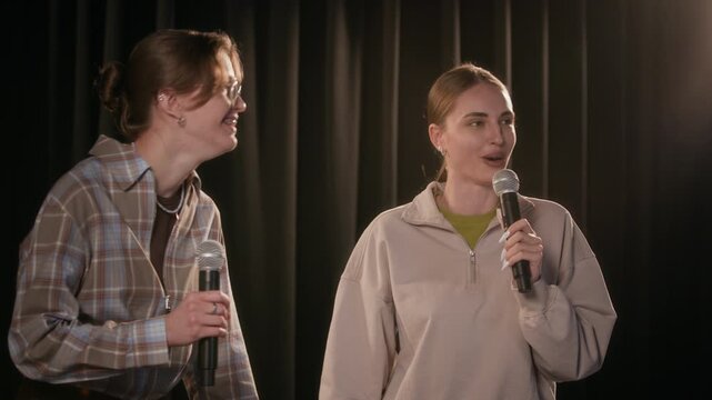 Medium shot of two laughing female comedians telling stories on stage against black curtain while performing back and forth comedy