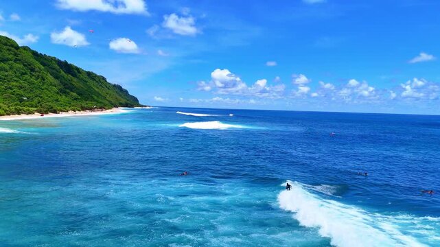 Aerial drone shot flying parallel to surfers riding waves near dramatic rocky cliffs in Bali, Indonesia. Dynamic action scene capturing the thrill of surfing in crystal clear ocean water