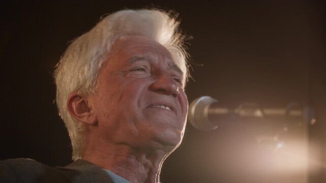 Close up shot of senior male musician singing song on stage under spotlight while performing live music during concert