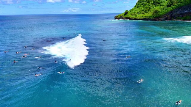 Aerial drone shot flying parallel to surfers riding waves near dramatic rocky cliffs in Bali, Indonesia. Dynamic action scene capturing the thrill of surfing in crystal clear ocean water