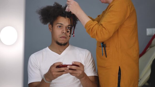 Young black man sitting in a salon chair while a hairdresser braids his hair into dreadlocks. Customer is looking at his phone