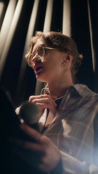 Vertical low angle shot of worried young adult female comedian with glasses fidgeting with fingers while checking speech script on clipboard standing behind curtain before show