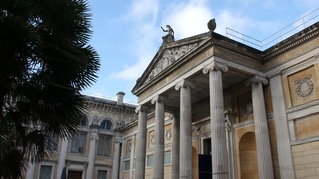 A neoclassical museum official stately building with tall stone columns and an ornate pediment, a statue sitting above the roof. Historic architecture rising over a courtyard in daylight