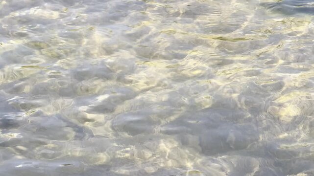 A mesmerizing close-up shot of crystal-clear water rippling over a shallow, sandy riverbed. Sunlight dances on the water's surface, creating shifting golden patterns and shimmering reflections. The ge