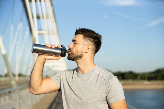 Young man staying hydrated after a workout, drinking fresh water from a reusable bottle on a sunny bridge