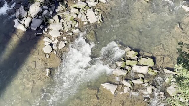 A top-down aerial-style shot of a crystal-clear mountain stream flowing over a rocky riverbed. The footage captures the dynamic movement of water as it cascades through rugged grey stones, creating wh