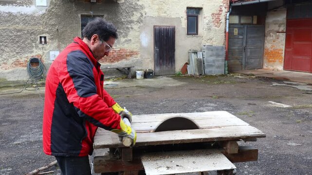 senior cutting wood into small pieces on a motorized circular saw in rural farm yard, performing manual preparation work for firewood storage as part of regular home activities in village environment