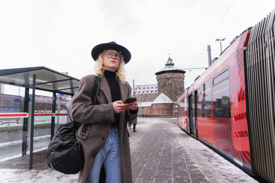 Young man waiting with smartphone at tram station in winter urban setting