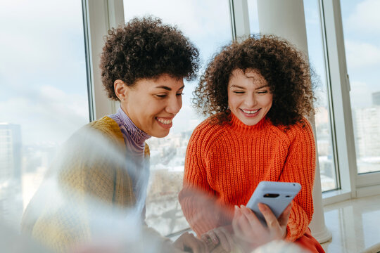 Young women with curly hair smiling and talking on windowsill indoors