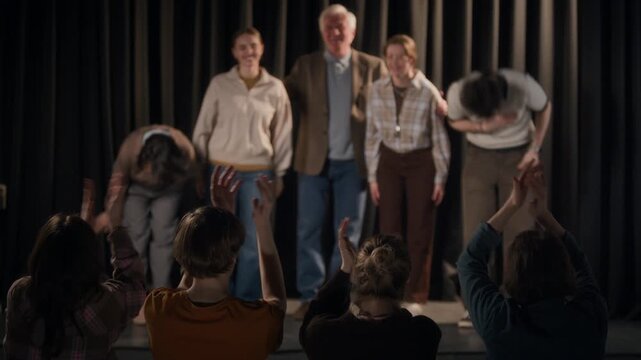 Rear view of unrecognizable people giving standing ovation to multigenerational crew of comedians standing on stage and bowing at end of stand up concert