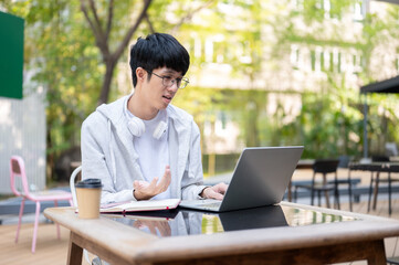 Fototapeta premium Glasses asian man student looking at laptop making confused hands as sitting at table in garden cafe