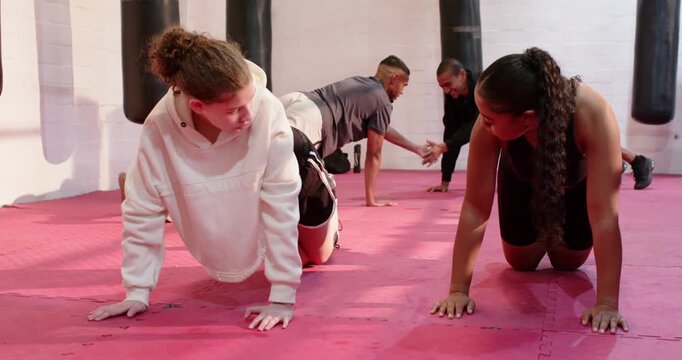 Diverse female workout partners shifting into and doing kneeling pushups on pink mat in boxing gym