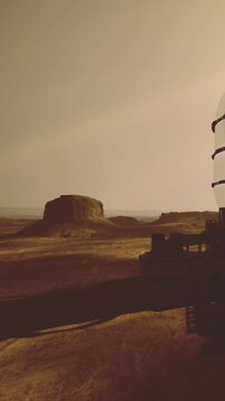 A rover sits on the Martian surface capturing the vast landscape. A distant rock formation towers under the light sky. The exploration is in a dry area with smooth terrain.