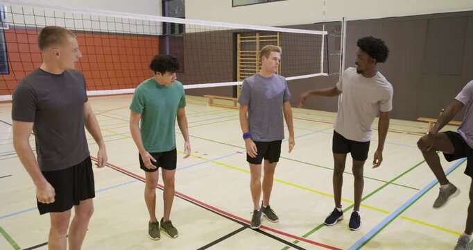 Diverse male athletes in gym performing knee holds and quad pulls by volleyball net before practice