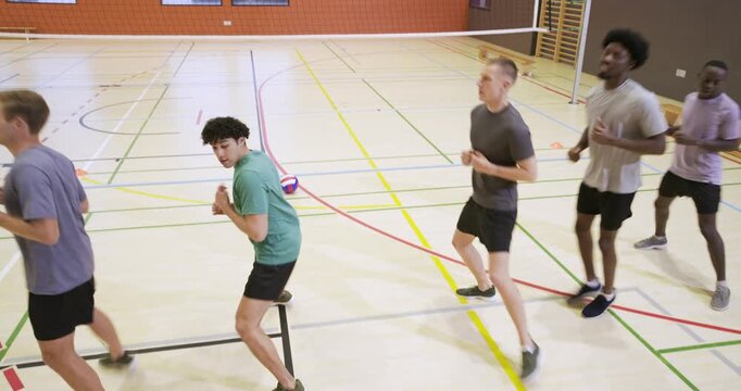 Five men warming up, jogging in line under volleyball net in athletic gear past two volleyballs