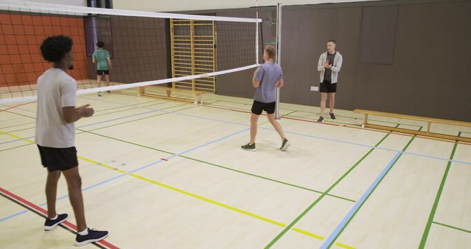 Diverse men playing as man in hoodie tossing purple volleyball over net, practicing passes in gym