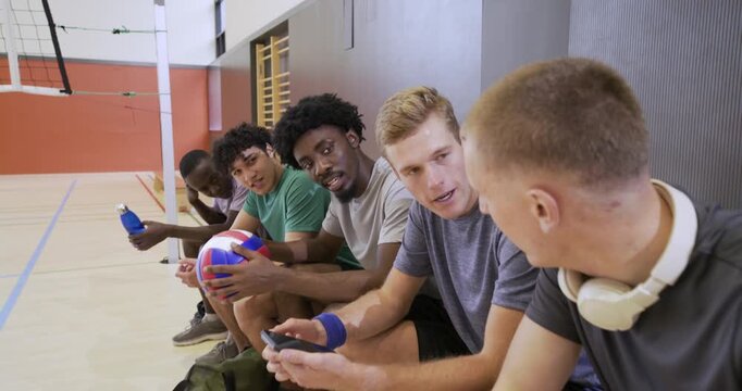 Diverse male teammates sitting on bench beside court, passing ball, sipping water, waiting for play