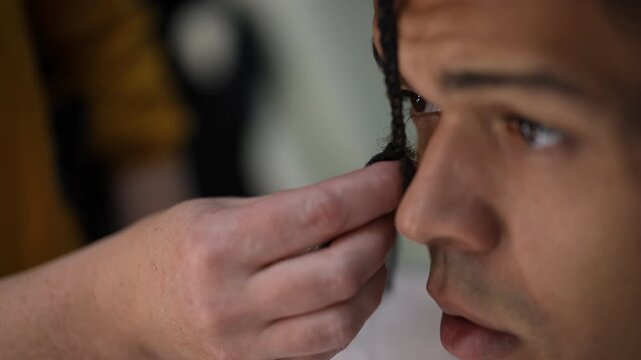 Close-up video of a hairdresser's hands skillfully plaiting a young man's dark hair. Professional hair styling service in a salon