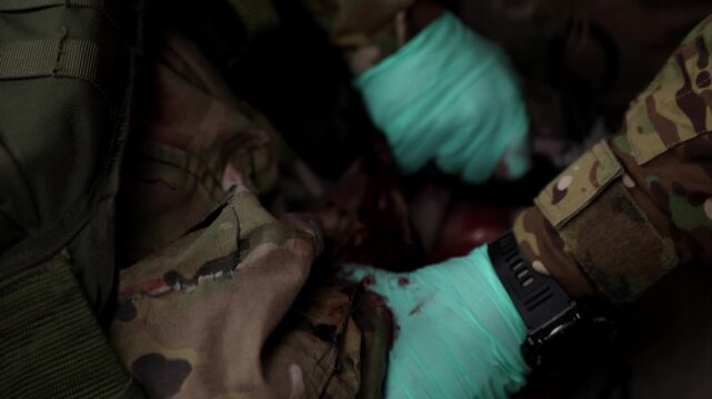 Close-up horizontal shot of a combat medic's hands in blue gloves tearing open a soldier's camouflage uniform sleeve to access and treat a bloody arm wound in the field