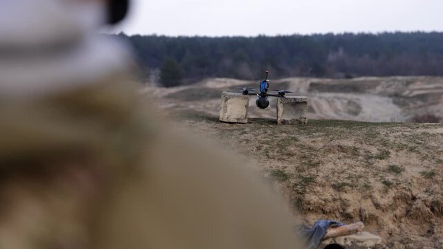 Wide horizontal shot of a tactical FPV drone with an explosive payload flying over a sandy training area, with a blurred soldier in the foreground