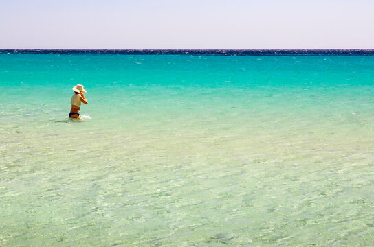 Travel, tourism: Woman wading in turquoise water at an tropical beach, wearing a hat, enjoying the summer vacation. Balistra beach, Corsica, France