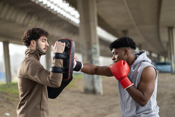 Two diverse males doing boxing training under a bridge. One man wearing red gloves is punching a...
