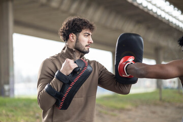 Male coach holds focus mitts as partner delivers a powerful punch during outdoor martial arts...