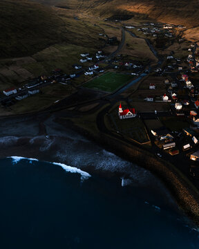 Aerial view of Sandavagur Church with its red roof and a green football pitch by the coast in Sandavagur, Vagar, Faroe Islands.