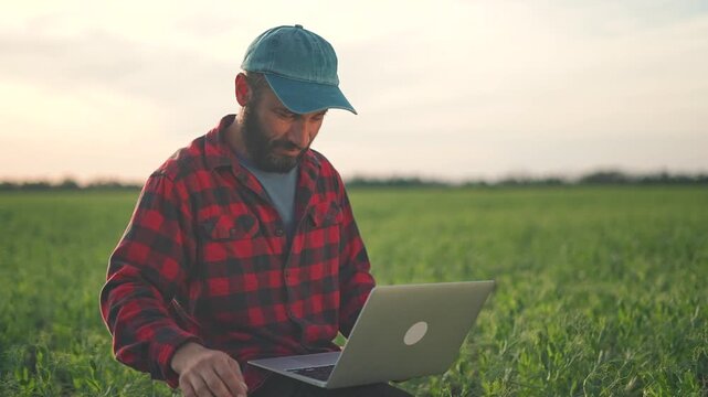 Farmer sits in field using laptop. Technology helps farmer check plant growth. Modern agriculture work computer. Farmer inspects plants and inputs data on laptop. Field work plant technology laptop.