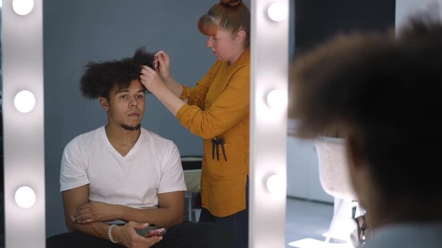 Young mixed race male model getting his afro hairstyle done by a female professional hairdresser in a backstage dressing room with a mirror
