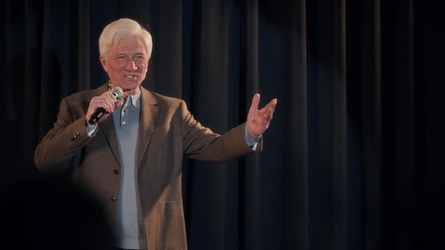 Medium full shot of senior male comedian walking on stage in spotlight beginning concert with crowd work asking spectator question during live performance on comedy club stage