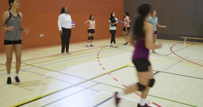 Diverse female team running rotations in school gym while coach tossing volleyball, practicing sets