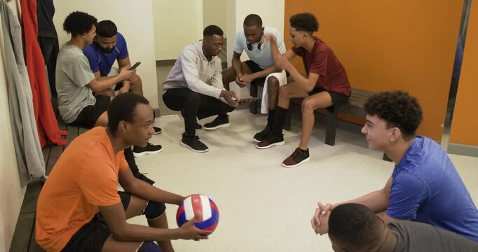 Diverse male team sitting benches lockerroom reviewing coach kneeling showing phone with volleyball