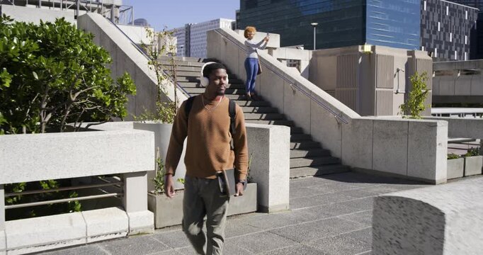 Diverse duo man walking to lens carrying skateboard while woman taking pictures with phone on stair