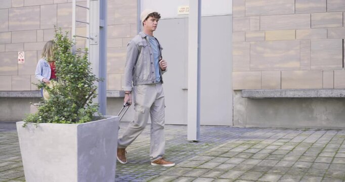 Two travelers walking across courtyard pulling suitcases as woman showing phone, man checking watch