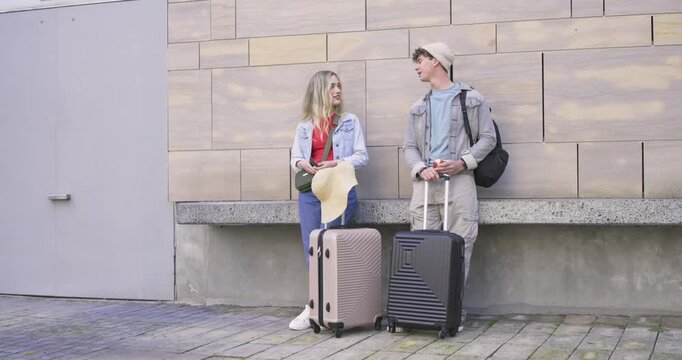 Two travelers with suitcases sitting on bench, woman checking phone, man approaching, sharing map