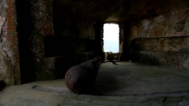 stone room inside Citadelle Laferri&egrave;re fortress in Haiti, featuring a large rusty cannon on the floor, arched window framing distant green valley under cloudy sky