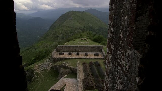 Rampart view from Citadelle Laferri&egrave;re fortress in Haiti, showing aligned stone cannon bases on grassy plateau, surrounded by verdant mountains and dramatic cloudy sky.