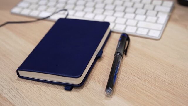 blue notebook with black pen beside a computer keyboard on a wooden desk indoors, featuring shallow focus and a clean workspace composition.