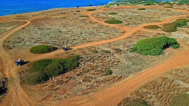 Drone view of quad bikes riding dusty tracks near Megaron des K&ouml;nigs and Potamos Beach by Agios Nikolaos, Greece. Coastal adventure scene with dry terrain, sea views and off road travel appeal.