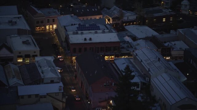 Footage of a small urban area at dusk, illuminated by streetlights and glowing interiors. The aerial view showcases diverse architectural styles with flat and gabled rooftops, some equipped