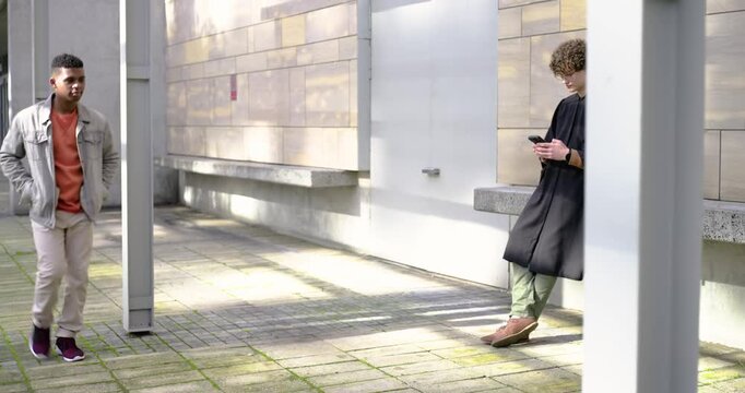 Adult gowned male grad leaning on seat at quad postgrad checking phone by cap or diploma, copy space