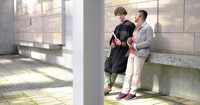 Two adult males leaning and talking after graduation at campus, graduate holding cap and diploma
