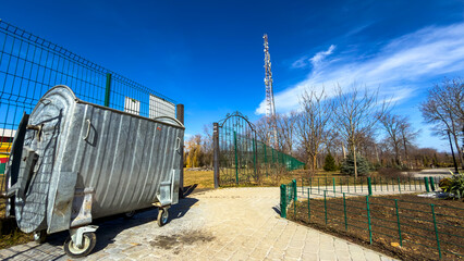 Fototapeta premium Park area with trash bin and cell tower. A trash bin stands near a park pathway with trees and a tall cell tower in the background on a sunny day.