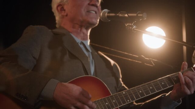 Tilt up shot of charismatic senior male comedy musician performing parody song on stage while playing acoustic guitar during live concert