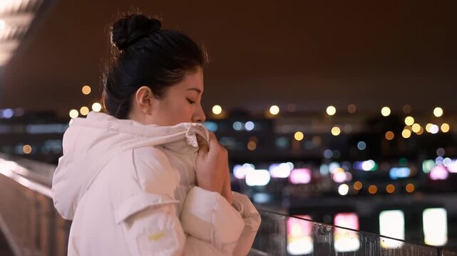 Pensive young woman in warm clothes at romantic dark balustrade over bright, observing the city lights, creating a quiet, melancholic and introspective atmosphere and cinematic winter moment