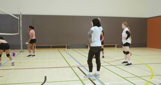 Diverse female volleyball team in sportswear training in gym, left player serving ball, diving
