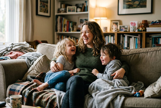 Woman sits on a beige couch with two children. She laughs with a young girl and boy on her lap. Bookshelves, framed photos, and a lamp fill the living room. Soft blankets and toys surround them