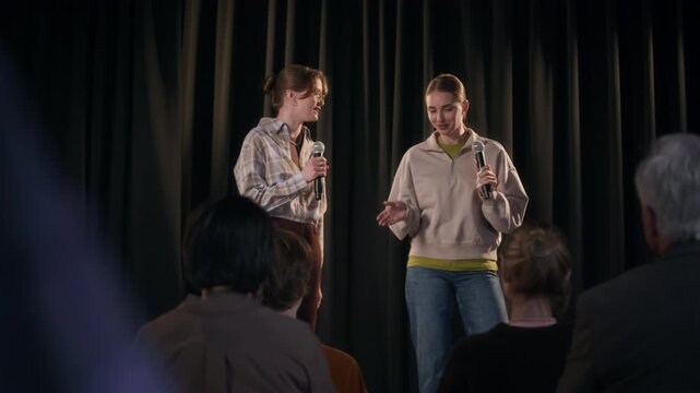 Shot from audience seats of two female comedians exchanging jokes in dialogue on stage during double act stand up performance at comedy club against black curtain